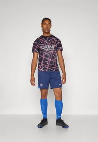 Noormees seisab seljas musta ja roosa mustriga PSG jalgpallivorm, tumesinised lühikesed püksid, sinised sokid ja mustad spordikepid, halli ühetoonilise tausta ees.