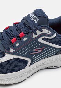 Zapatilla deportiva navy con parte superior de malla, acentos rojos y cordones. Cuenta con una suela resistente y detalles texturizados a los lados. Branding visible.