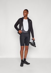 Svart sykkeljakke med lange ermer over en hvit skjorte, kombinert med svarte bib-shorts. Modellen holder en svart hjelm og har på seg sykkelsko.