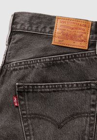 Gros plan sur un jean en denim Levi's gris foncé montrant la poche arrière, l'étiquette rouge Levi's et un patch en cuir fauve avec logo et texte.