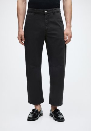 Mann iført svarte avkuttede bukser og svarte skinnloafers, stående mot en lys bakgrunn.