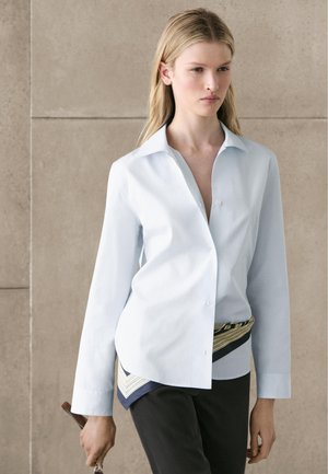 Blondynka w jasnoniebieskiej koszuli na guziki i czarnych spodniach, trzymająca torebkę na brązowym pasku, stojąca na tle beżowej ściany.