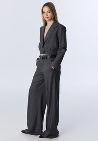 Grijze cropped blazer met een getailleerde taille en grote knopen, gecombineerd met brede grijze broekspijpen. Soepele stof met een lichte glans en zakken.