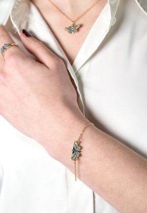 Gouden armband met een vlindermotief en blauwe en groene accenten, bevestigd aan een delicate ketting, rustend op de pols van een persoon.