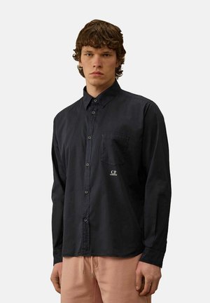 Camicia nera con bottoni e maniche lunghe, taschino sul petto e dettaglio del logo. Indossata con pantaloni rosa chiaro, con una texture liscia.