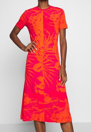 Kobieta ubrana w sukienkę z krótkim rękawem do kolan w jasny czerwono-pomarańczowy wzór kwiatowy, pozująca z jedną ręką na biodrze.