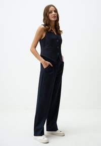 Navyblauwe vest met drie knopen en twee zakken, gecombineerd met bijpassende brede broek. Gemaakt van een gladde, gestructureerde stof.