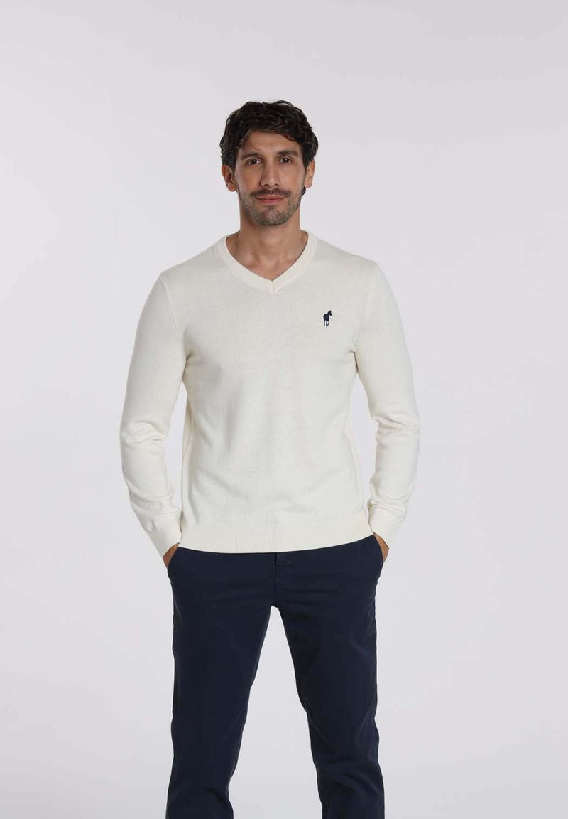 Pull léger crème en v en tissu doux, avec un logo marine sur la poitrine et des poignets côtelés. Porté avec un pantalon marine.