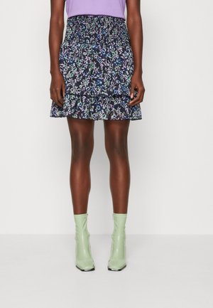 Vrouw draagt een korte rok met bloemmotief en een geplooide taille, een lavendelkleurige top en lichtgroene enkellaarsjes, staand tegen een witte achtergrond.
