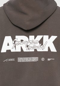 Szürke kapucnis pulóver nagy fehér "ARKK" logóval, alatta a "Innovation Included" felirat stilizált betűtípussal. Egyszerű dizájn, puha textúra.