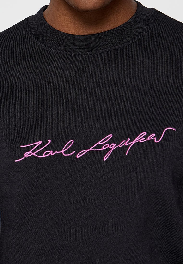 Nahaufnahme eines schwarzen Strickpullovers mit rosa, geschwungener Stickerei, die "Karl Lagerfeld" auf der Brust zeigt.