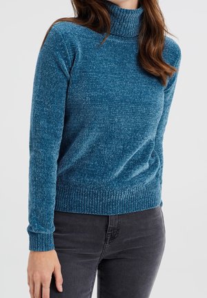 Femme portant un pull à col roulé côtelé bleu sarcelle et un jean gris foncé, debout devant un fond clair uni.