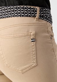Beige housut, joissa on tekstuuripinta, mustalla kuvioidulla vyötärönauhalla ja takatasku raidoitettuna yksityiskohtana.