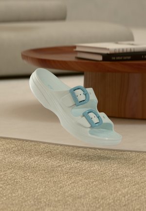 Sandalo a ciabatta azzurro chiaro con due fibbie regolabili che fluttua sopra un tappeto intrecciato, un tavolino in legno e un divano sfocato sullo sfondo.