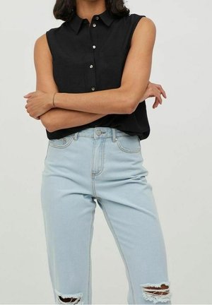 Mouwloos zwart overhemd met knopen, gecombineerd met lichtblauwe gescheurde jeans, gedragen door een persoon die met de armen gekruist staat.