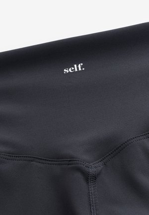 Material negru văzut de aproape cu text alb "self." imprimat lângă cusătura cusută pe un material neted și elastic, probabil îmbrăcăminte sport.