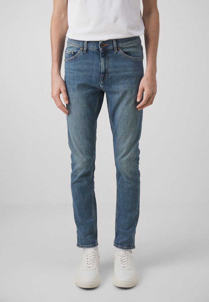 Jean en denim bleu avec une coupe slim, délavage, cinq poches et couture contrastée, associés à des baskets blanches.