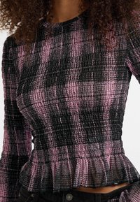 Frau trägt ein enges, langärmliges Oberteil in einem rosa und schwarzen karierten Muster mit einem Rüschensaum, kombiniert mit schwarzen Hosen.