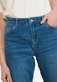 Gaiši zili džinsa bikses ar augstu jostasvietu, metāla pogas aizdari un piecām kabatām. Audumam ir gluda tekstūra un viegla izbalēšana.