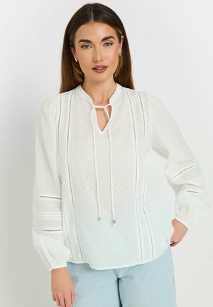Hvid bluse med en nøglehulslukning og snor. Har lodrette striber, strukturerede polka prikket og lange, puffede ærmer.
