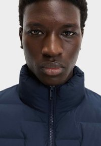 Chaqueta de plumas azul marino con alto cuello y cremallera completa, que presenta una textura suave y costuras horizontales a lo largo del cuerpo.