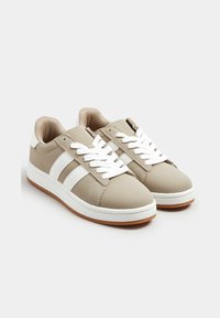 Jasno beżowe sneakersy wykonane z materiału syntetycznego, z białymi paskami, okrągłym noskiem, białymi sznurowadłami, wyściełanym kołnierzem oraz gumową podeszwą.