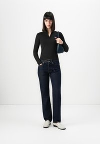 Vrouw met zwart steil haar staat in een zwart langemouwen top, donkerblauwe jeans, witte schoenen en houdt een marineblauwe schoudertas vast.