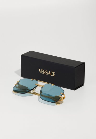 Occhiali da sole Versace con lenti blu scure, montatura in metallo dorato e accenti decorativi, esposti accanto a una scatola nera con logo.