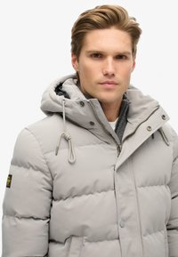 Grå pufferjakke med aftagelig hætte, med lynlås, justerbar snøre og ribstrikkede manchetter. Struktureret, kviltet mønster.