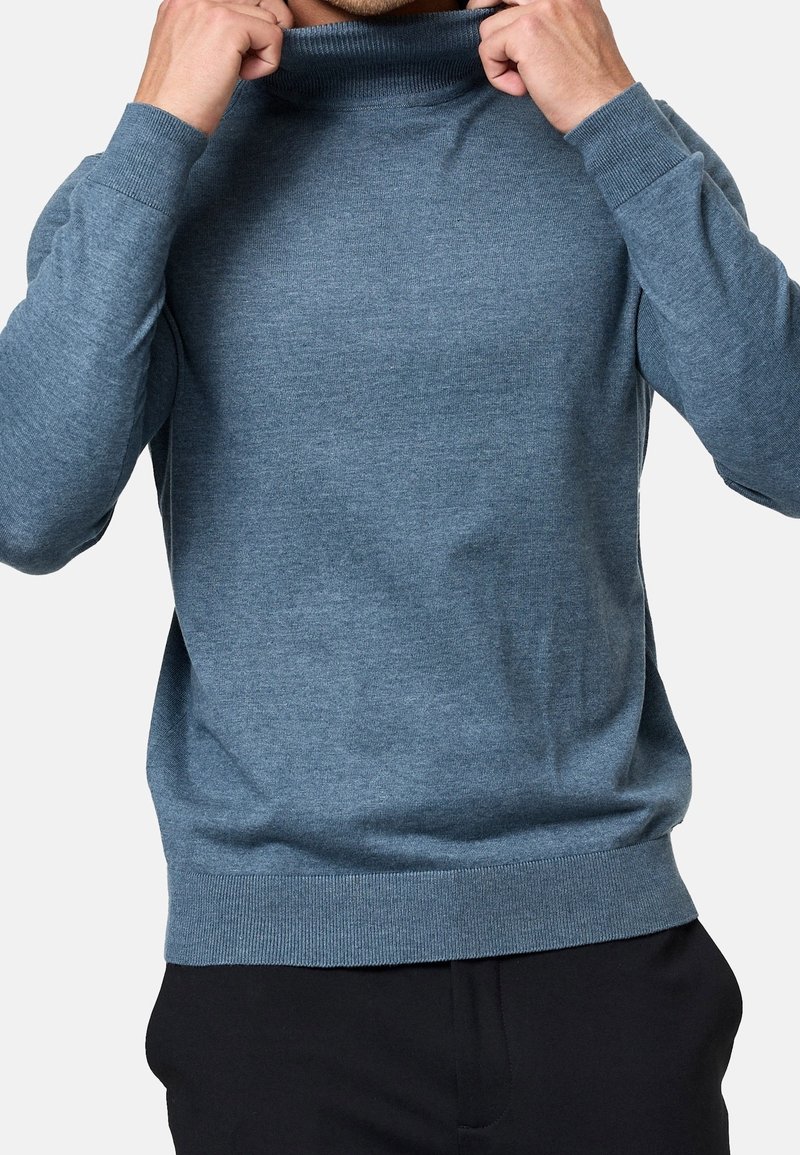 Pull en tricot bleu avec un col montant, des poignets et une taille côtelés. Texture lisse et design ajusté, partiellement rentré.