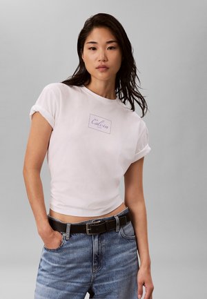 Młoda kobieta o ciemnych włosach nosi biały krótki T-shirt Calvin Klein i niebieskie jeansy z czarnym paskiem, pozując na szarym tle.