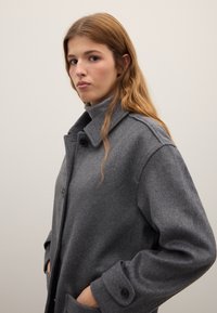 Grauer Wollmantel mit einem Kragen, Knopfleiste und Seitentaschen. Der Stoff hat eine weiche Textur und eine lockere Passform.