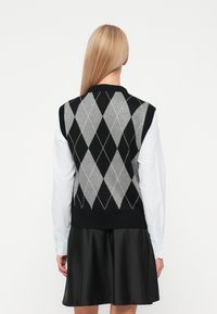 Fekete és szürke rombuszmintás kötött mellény bordás nyakkivágással, fehér hosszúujjú ing fölé húzva, fekete szoknyával párosítva.