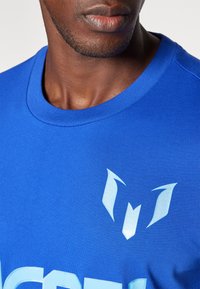 Blauses Baumwoll-T-Shirt mit Rundhalsausschnitt, ausgestattet mit hellblauen Grafiken: einem stilisierten Logo und fettem Text. Glatte Textur.