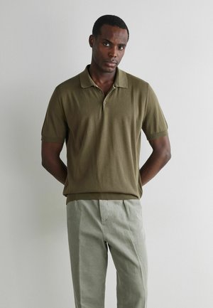 Mann steht vor weißer Wand, trägt ein olivgrünes kurzärmeliges Polohemd und hellgraue Hose, die Hände hinter dem Rücken, schaut nach vorne.