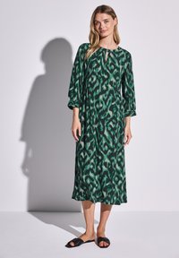 Vihreä puolipitkä mekko, jossa on avainaukko kaula-aukko, 3/4-hihat ja kuvioitu rakenne. Yhdistetty mustiin tasapohjaisiin sandaaleihin.