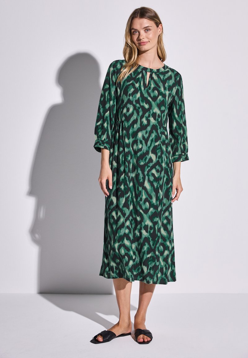 Vihreä puolipitkä mekko, jossa on avainaukko kaula-aukko, 3/4-hihat ja kuvioitu rakenne. Yhdistetty mustiin tasapohjaisiin sandaaleihin.