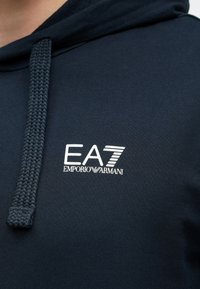 Sötétkék kapucnis pulóver puha anyagból, fonott húzózsinórral és fehér EA7 Emporio Armani logóval a mellrészen.