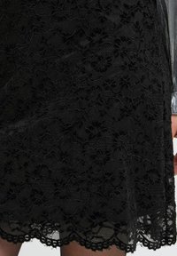 Zwart kanten jurk met bloemenpatronen, voorzien van een gegolfde zoom en gestrikte lagen. Het materiaal lijkt lichtgewicht en iets doorzichtig te zijn.