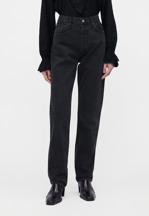Femme portant un jean noir taille haute associé à des bottines noires à talons et un chemisier noir à manches longues avec des poignets à volants.