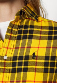 Chemise à carreaux jaune et noir avec des accents rouges, boutonnée au col, avec un petit logo brodé représentant un joueur de polo noir.