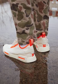 Adidas NMD kengät valkoisina, joissa on oranssit yksityiskohdat, pehmustettu pohja, teksturoitu sivu ja heijastavat yksityiskohdat, yhdistettynä kamoflammipöksyihin.