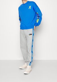 Ljusblå sweatshirt med rund halsringning, med gröna och gula logotryck. Grå sweatpants med blå sidostråk och broderad detalj.