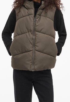 Person iført en brun polstret ærmeløs vest over en sort langærmet sweater, hænderne i vestens lommer og sorte bukser.