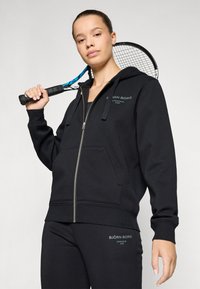 Hoodie preto com fecho éclair, feito de tecido suave, apresentando um bolso frontal, capuz com cordão e texto impresso em verde. Combinado com calças a condizer.
