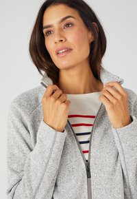 Felpa grigia con zip e un finish texturizzato, con colletto alto. Sotto si intravede una camicia crema con righe orizzontali rosse e blu.