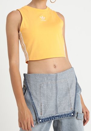 Yellow crop top with round neckline, white Adidas logo, and white side stripes. Worn with light blue denim overall-style pants featuring front pockets.