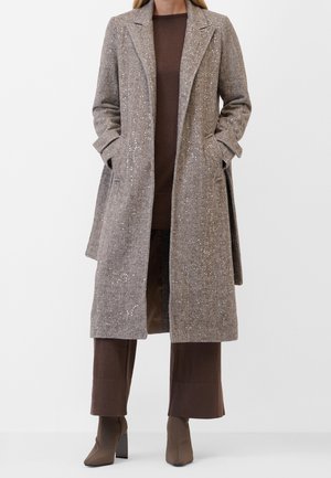 Femme portant un long manteau beige texturé sur une robe marron et un pantalon large, associé à des bottines taupe à talons blocs.