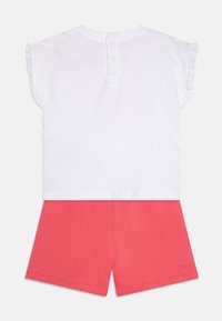 Weißes Kurzarmoberteil mit Rüschensaum und drei Knöpfen auf der Rückseite, kombiniert mit korallenroten Shorts. Aus weichem Baumwollmaterial gefertigt.
