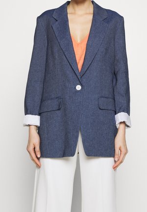 Personne portant un blazer bleu foncé avec des poignets blancs retroussés, un haut orange à col en V et un pantalon blanc, visible des épaules jusqu'à mi-cuisse.